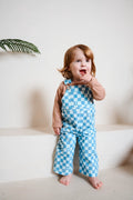 A toddler standing barefoot on a white step, wearing a brown long-sleeve shirt and light blue and white checkered overalls, with a green palm leaf visible in the upper left corner.